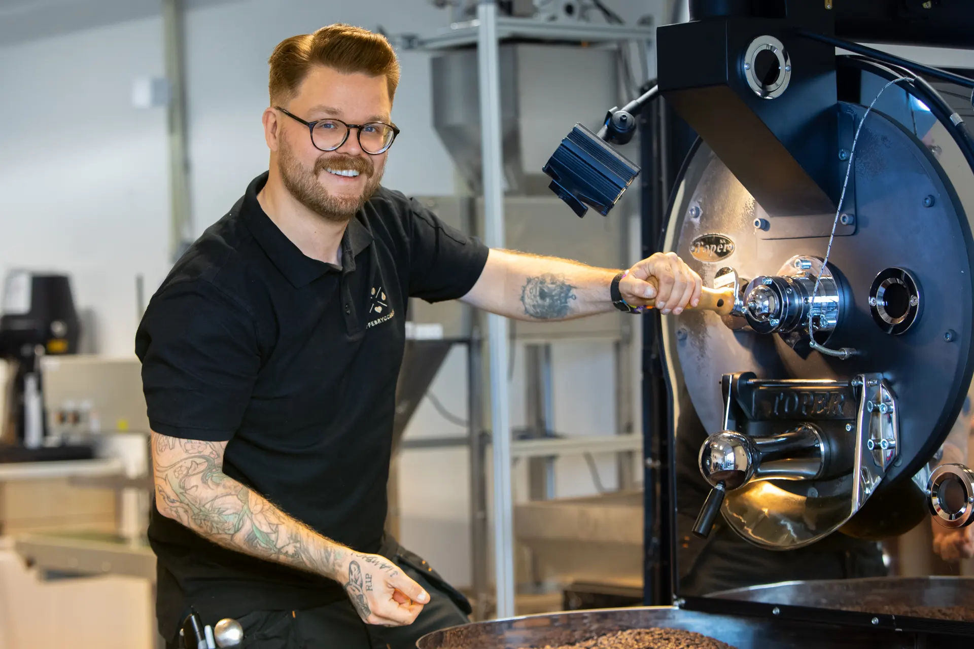 Mann med briller og tatoveringer som betjener en stor kommersiell kaffebrenner og viser frem førsteklasses kaffemaskin bedriftskompetanse i et moderne anlegg.
