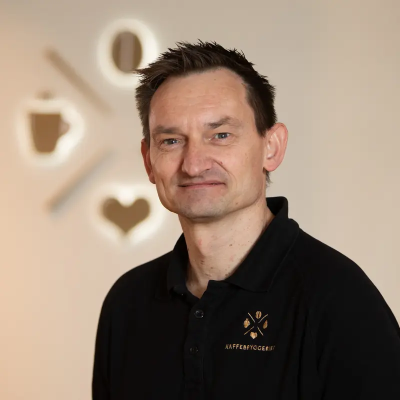 Smilende middelaldrende mann, iført svart poloskjorte med Organisasjon Kaffebryggeriet-logo, stående foran en sløret veggkunst med kaffetema.