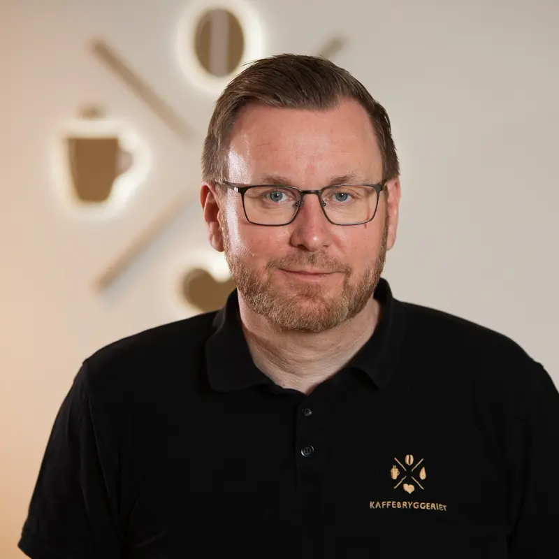 En mann med briller og skjegg, iført svart polo med "KAFFEBRYGGERIET"-logo, står innendørs med et uskarpt veggmotiv med kaffetema i bakgrunnen.