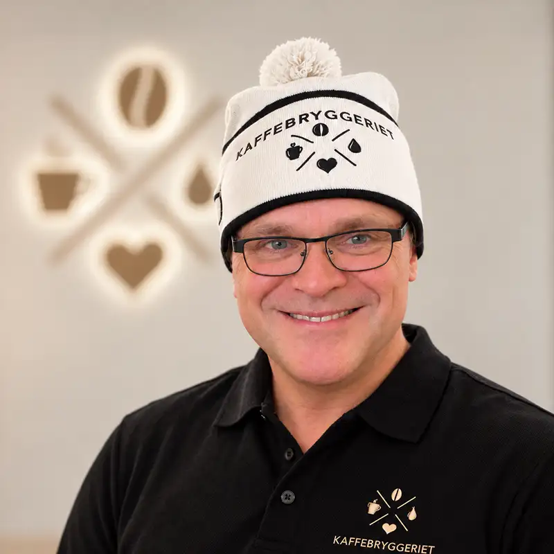 En smilende mann har på seg en hvit lue og en svart polotrøye, begge med Kaffebryggeriet-logoen.