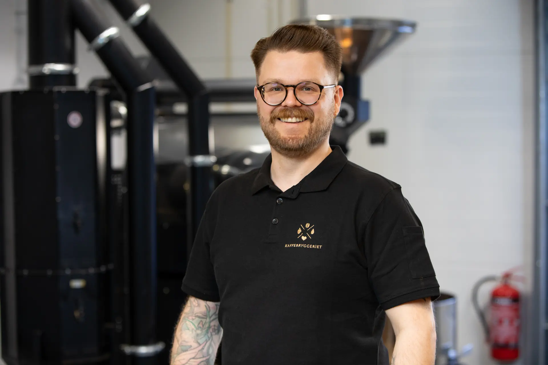 En mann med briller og tatoveringer, iført svart polo med kaffebrenneri-logo, står smilende i et industrielt miljø og tilbyr stolt kaffeløsning til bedrift blant kaffebrenneriutstyr i bakgrunnen.
