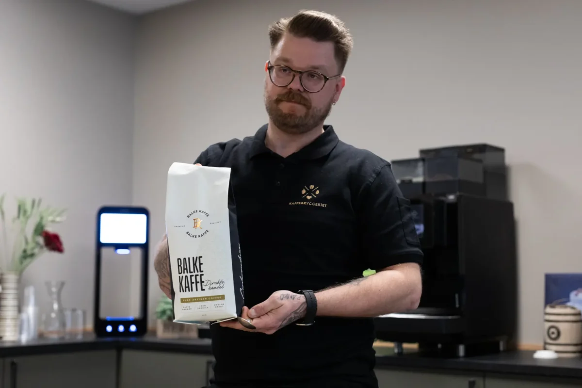 En mann i svart poloskjorte holder en pose med Balke Kaffe i et moderne kjøkken, som viser en ideell kaffeløsning for bedrift med kaffemaskin og andre gjenstander i bakgrunnen.