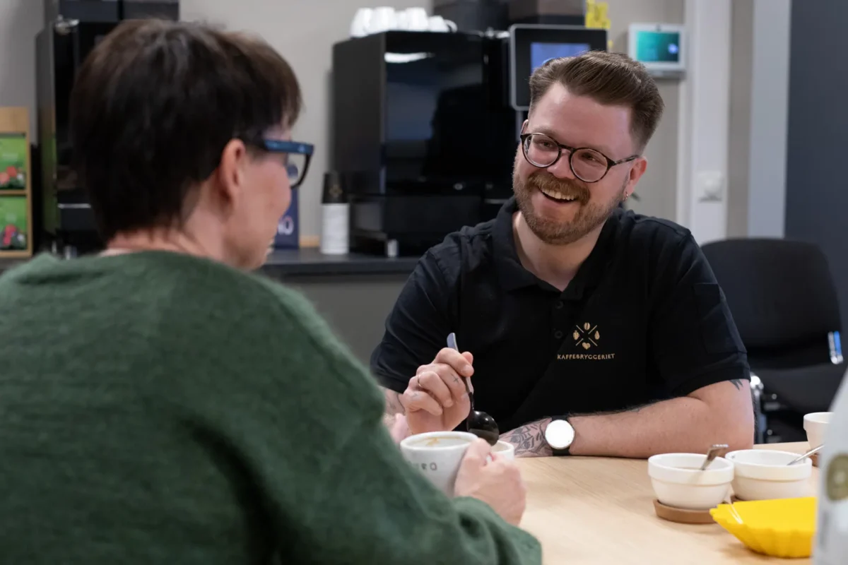 To personer sitter ved et bord og snakker sammen over en kaffe i et moderne kjøkken, mens de nyter kaffeløsningen til bedrift. Den ene personen smiler og holder i en skje, noe som skaper en avslappet og innbydende atmosfære.
