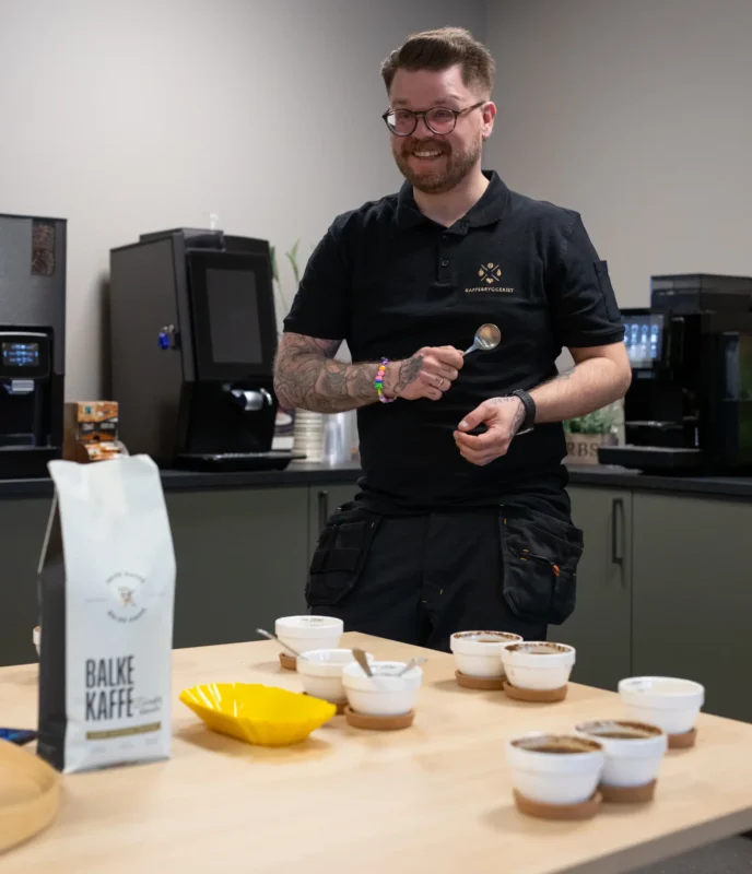 En person står smilende med en skje i hånden i en kjøkkenkrok, med kaffekopper og en pose merket "BALKE KAFFE" på bordet - og viser frem en ideell kaffeløsning til bedrift.