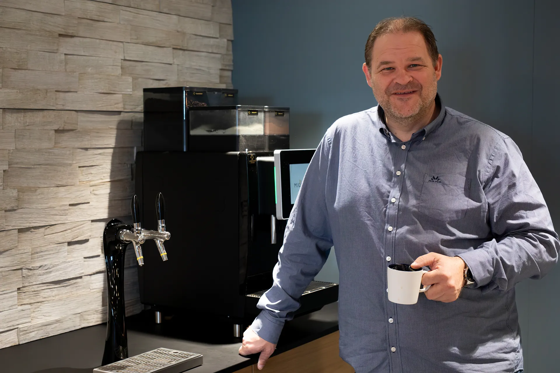 En mann i blå skjorte står ved en kaffemaskin, holder et hvitt krus og smiler, og viser frem en kaffeløsning for bedrift i et moderne kjøkken- eller pauseromsmiljø.