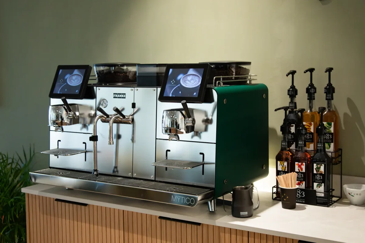 En moderne Mytico kaffemaskin med doble dispensere og digitale skjermer sitter på benkeplaten, ledsaget av smakssirupsflasker og trerørere.