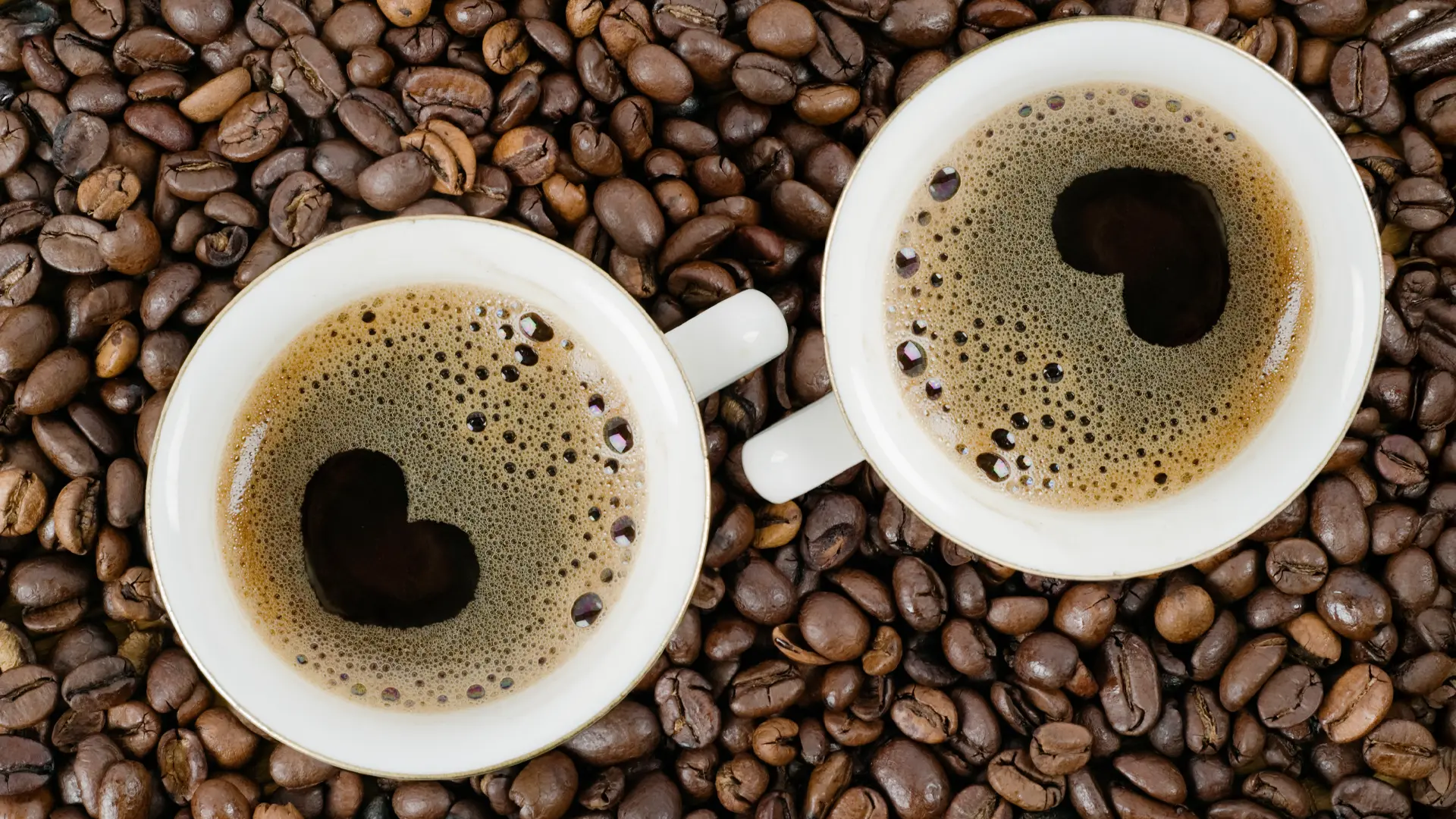 To hvite kaffekopper fylt med svart kaffe fra en ny kaffemaskin står på en seng av ristede bønner, og hver kopp har et hjerteformet mønster i cremaen.