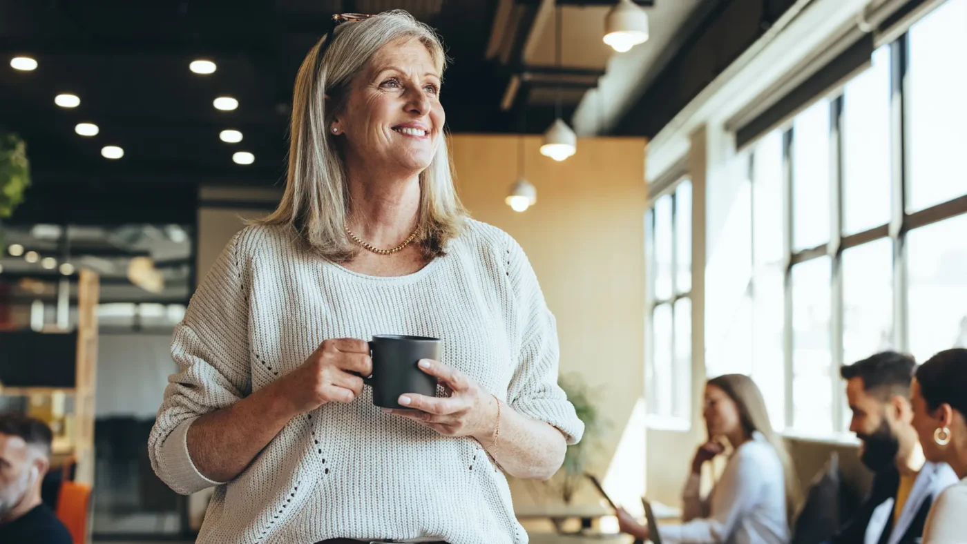 En eldre kvinne med et krus i hånden står og smiler ved kaffemaskinen i et moderne kontorlokale, mens flere personer diskuterer i bakgrunnen.