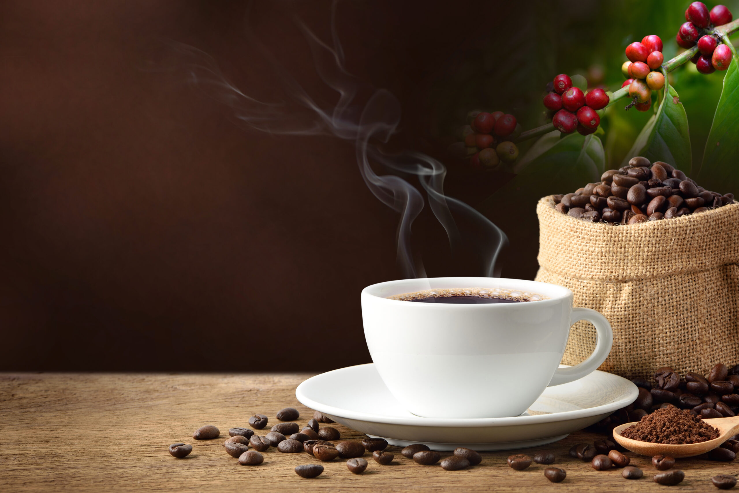 En rykende kopp kaffe på en tallerken ved siden av en burlapsekk som flommer over av miljøvennlige kaffebønner, med røde kaffekirsebær og en treskje med malt kaffe i nærheten.