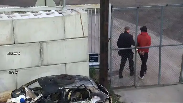To personer står ved et inngjerdet gjerde, den ene i svart dress og den andre i rød jakke og svarte bukser, og legemliggjør sikkerhet på stedet mens de oppmerksomt kartlegger betongområdet med en utbrent bil i forgrunnen.