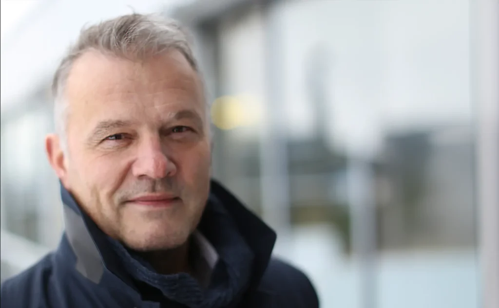 En middelaldrende mann med kort grått hår og iført mørk jakke står foran en uskarp bakgrunn. Velkommen til vår nye daglige leder!