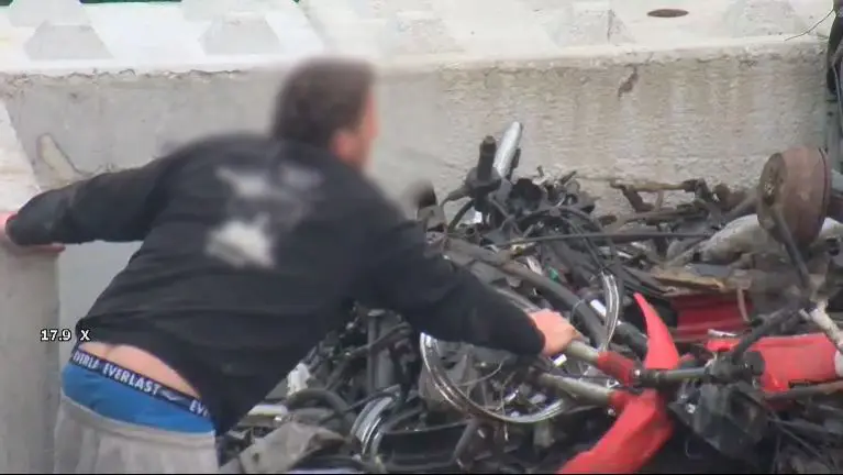 En person iført svart skjorte og blå linning trekker en rød sykkelramme fra en haug med skrapmetall mens han lener seg mot en betongbarriere, som minner om nylige nyheter hvor en rask reaksjon hindrer tyveri – bevæpnet mann pågrepet hos bilopphugger i Arendal.