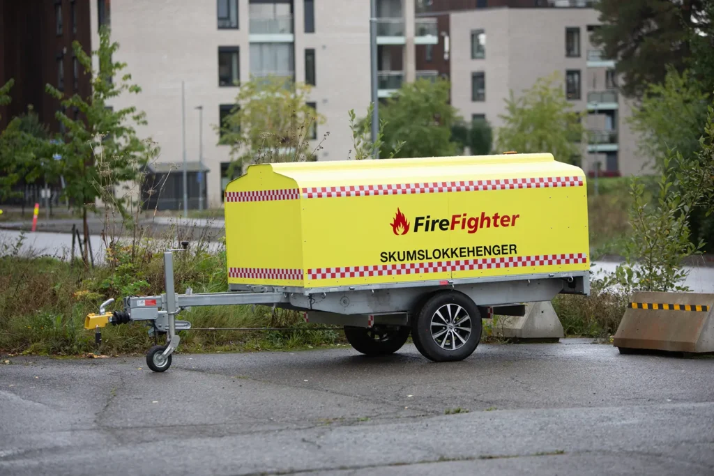 En gul slokkeskumhenger merket «Brannmann Skumslokkehenger» står parkert i et asfaltert, delvis gjengrodd område nær bebyggelse, klargjort for effektiv brannslukking for avfallsbransjen.